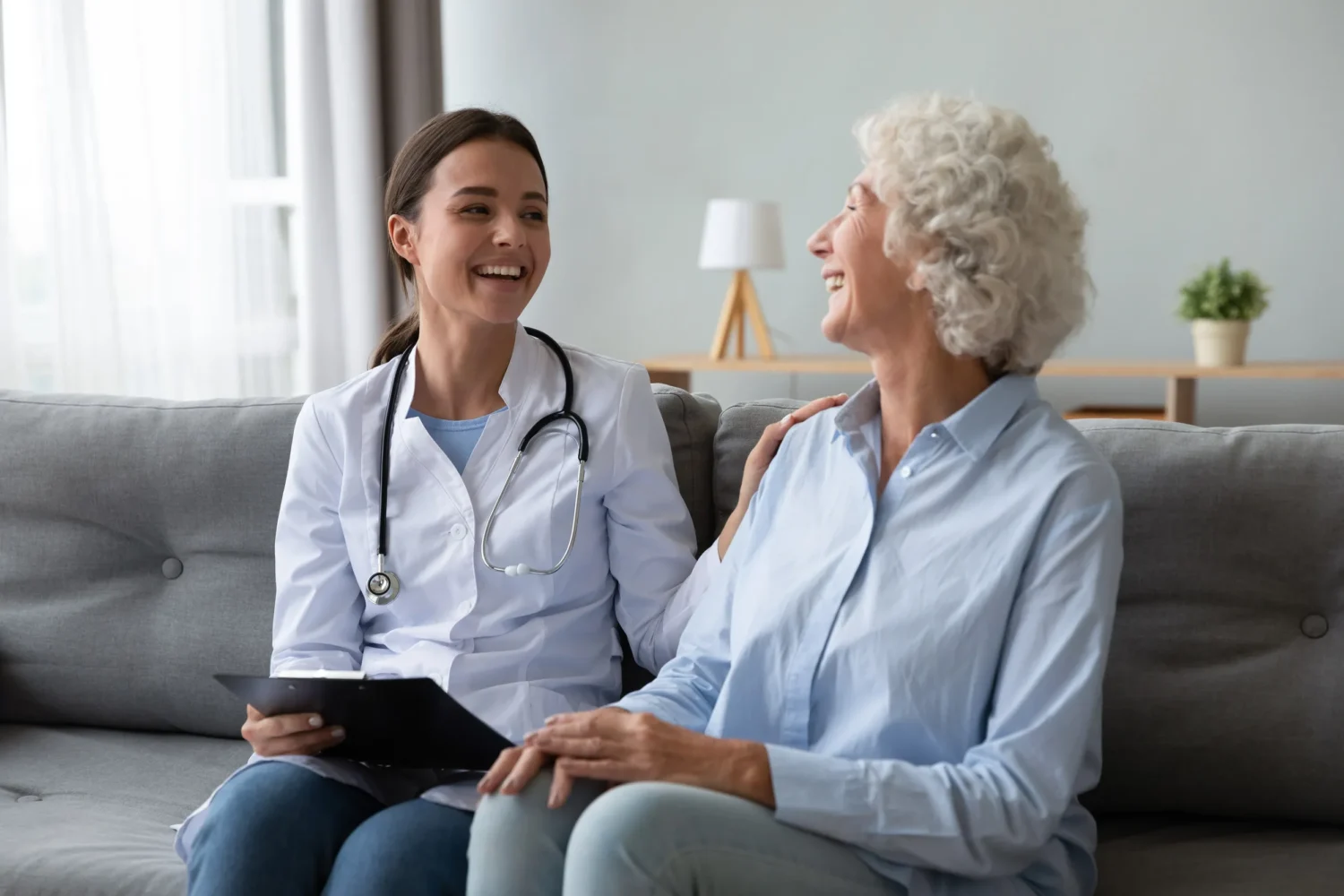 Friendly young nurse in white coat holding clipboard laughing with elderly female patient during homecare visit consultation