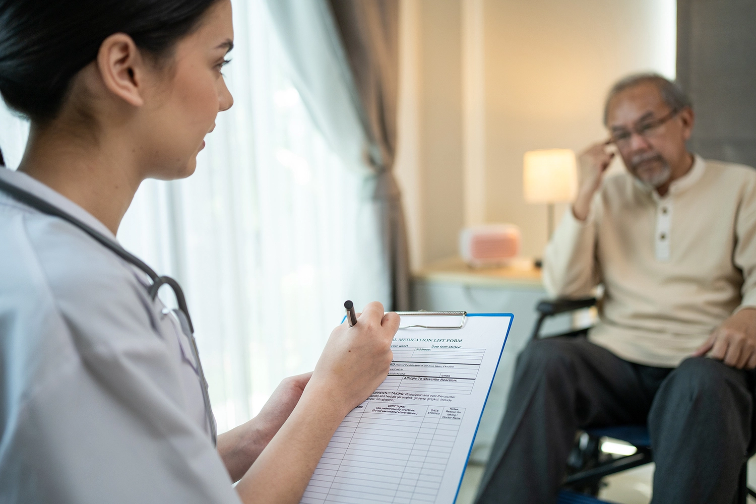 A healthcare professional takes notes on a clipboard while interacting with an elderly patient in a well-lit room