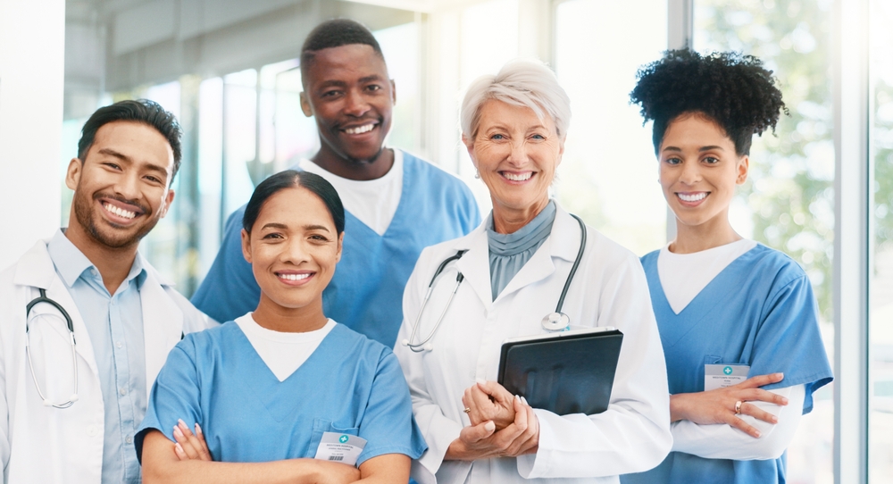 A diverse group of healthcare professionals stands together, wearing scrubs and lab coats, with warm smiles in a bright hospital setting.