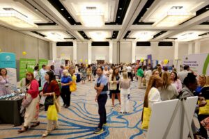 A bustling conference hall filled with attendees exploring exhibition booths, surrounded by bright banners and vibrant decor.