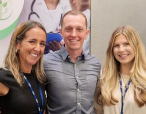 Three individuals pose together at an event, smiling, with a healthcare-themed backdrop behind them