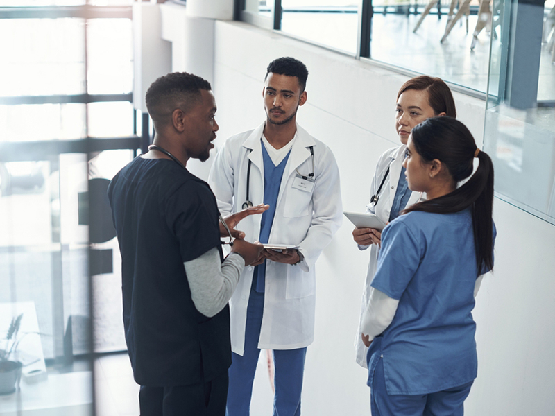 A diverse group of healthcare professionals stands together, wearing scrubs and lab coats, with warm smiles in a bright hospital setting.